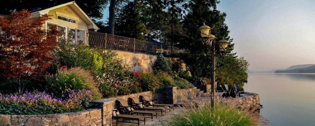 Before and after of a backyard transformed from an empty lawn into a luxurious poolside pavilion with stone columns, orange sectional, pergola, and elegant garden accents.