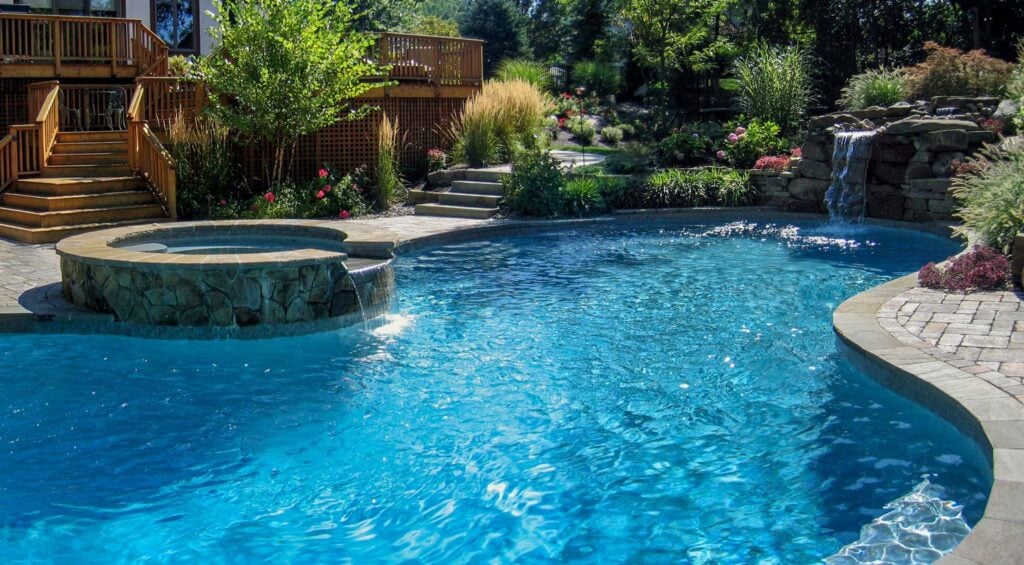 Before and after of a backyard space transformed from a plain deck into a vibrant garden retreat with a koi pond, stone path, and wooden seating area with dogs overlooking the view.