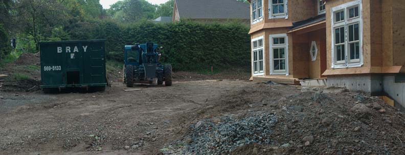 Front yard under construction with exposed soil, building materials, a green dumpster, and unfinished home exterior