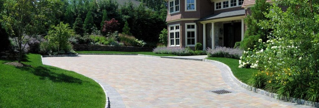 Professionally designed driveway with paver stones, manicured shrubs, lush lawn, and vibrant garden beds in front of a luxury home