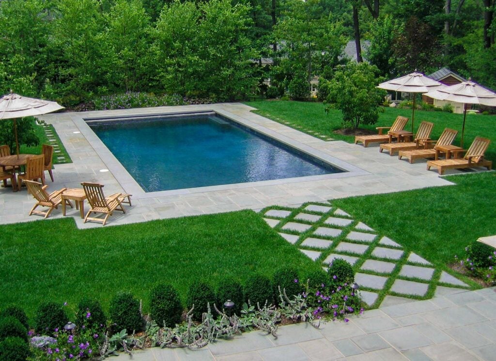 Before and after of a backyard renovation showing a transformation from plain lawn to a luxurious pergola-covered lounge with outdoor kitchen, poolside seating, and vibrant landscaping.