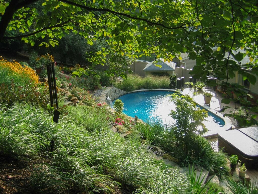 Before and after transformation of a sloped backyard into a structured outdoor living space with terraced stone walls, plantings, and a staircase by CLC Landscape Design.