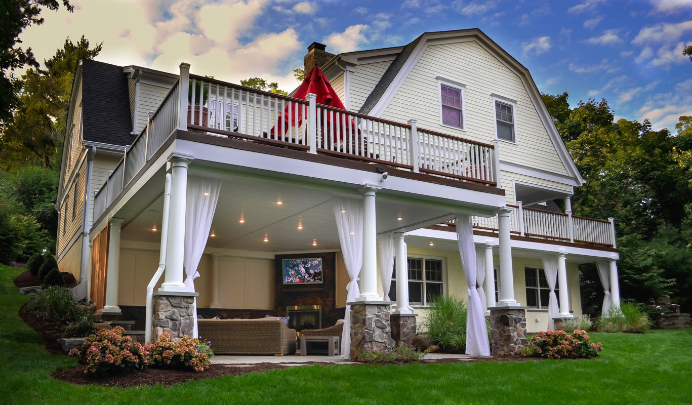 Two-story home in Chatham, NJ with upper deck and covered patio lounge below, featuring white railings, columns, and string lighting.