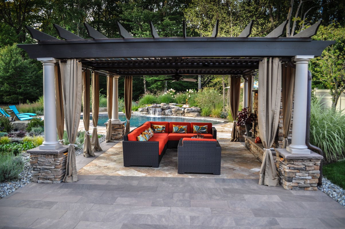 Outdoor poolside pavilion with stone pillars, taupe curtains, and a bold orange sectional by CLC Landscape Design in New Jersey.