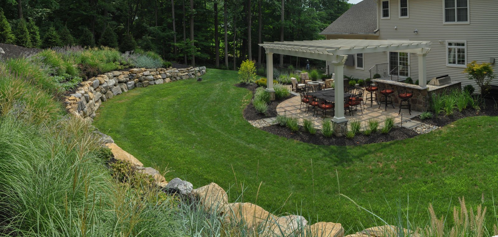 Expansive backyard featuring a white pergola over an outdoor dining and kitchen area with bar seating, surrounded by manicured lawns, stone retaining walls, ornamental grasses, and layered plantings.