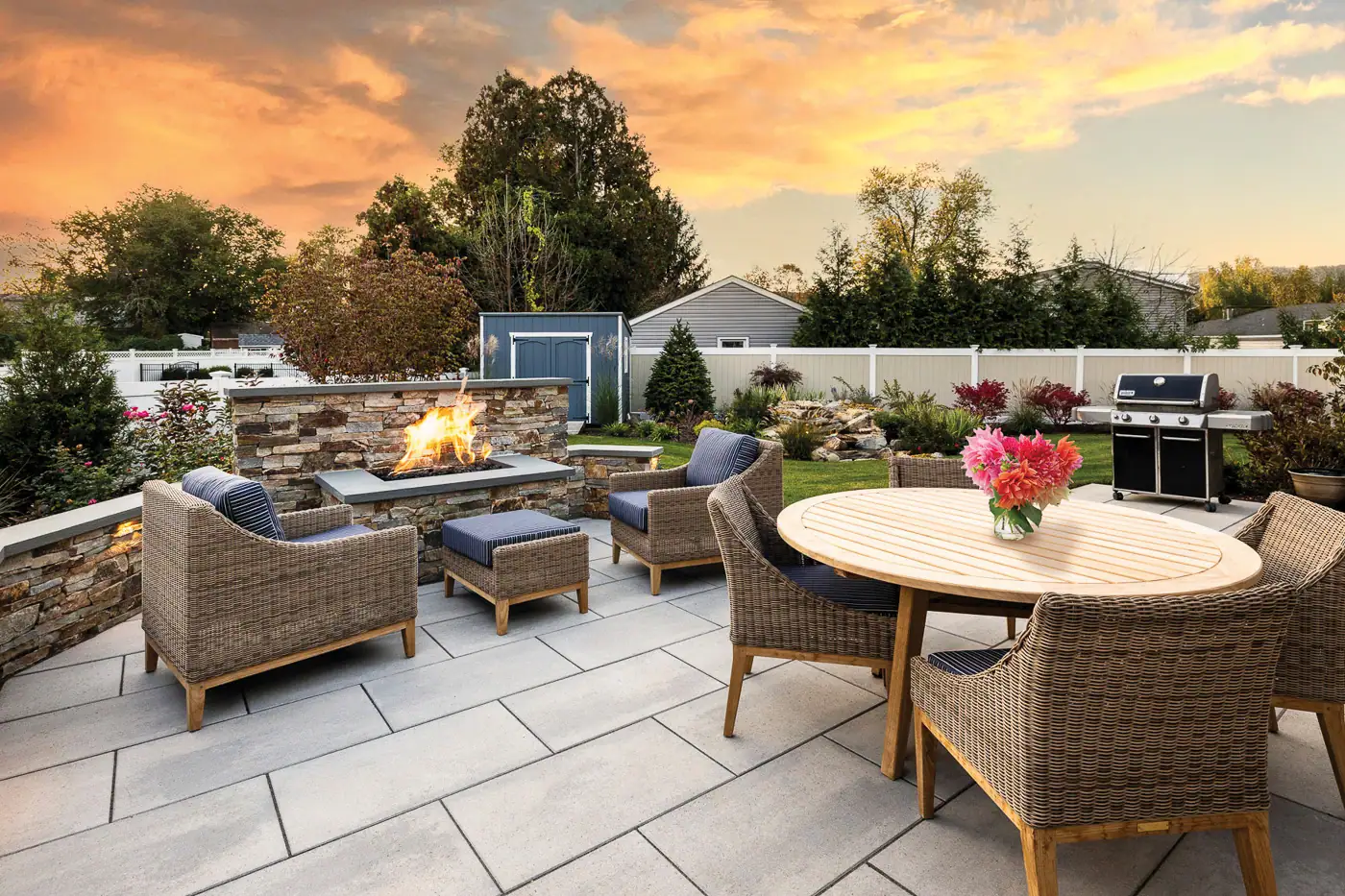 Modern paver patio by CLC Landscape Design in Pompton Lakes, NJ — cozy seating arranged around a custom fire pit within a stone seat wall, with a subtle pondless water feature, surrounded by lush plantings and soft evening light