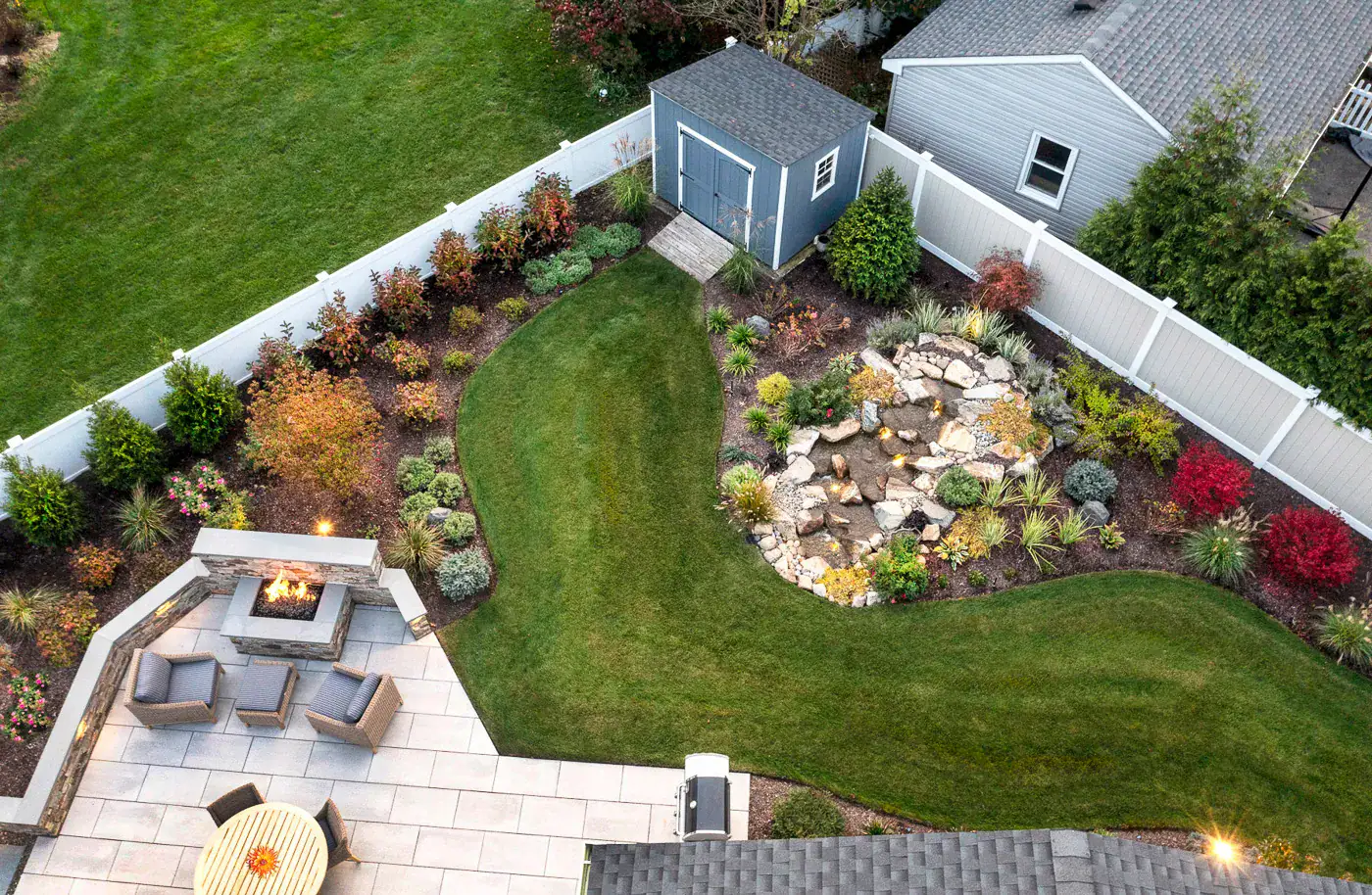 Aerial view of a modern backyard by CLC Landscape Design in New Jersey—elegant paver patio with built-in fire pit and cozy seating area, flowing into a lush lawn bordered by a naturalistic pondless water feature with stones and ornamental plantings.