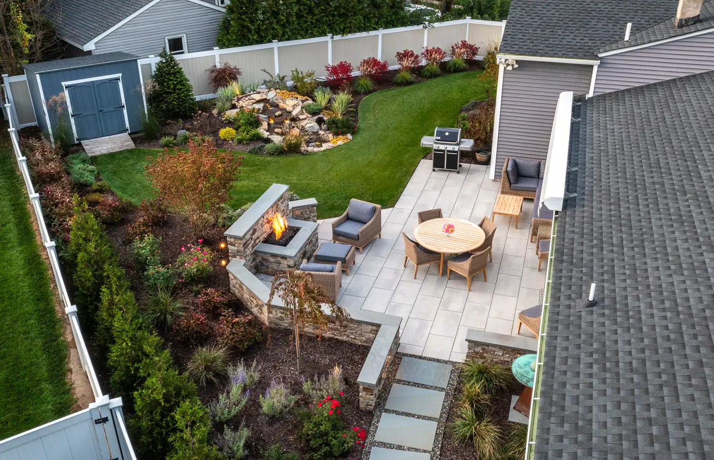 Modern patio with built-in fire pit by CLC Landscape Design in Pompton Lakes, NJ—stone pavers, intimate seating area, and a cascading pondless water feature framed by vibrant plantings.