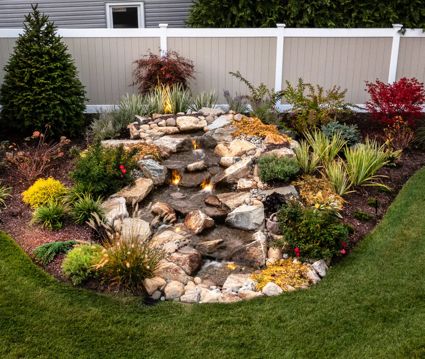 Custom pondless waterfall with integrated fire features by CLC Landscape Design in New Jersey—natural boulders and layered plantings create a dramatic fire-and-water focal point for this residential backyard.