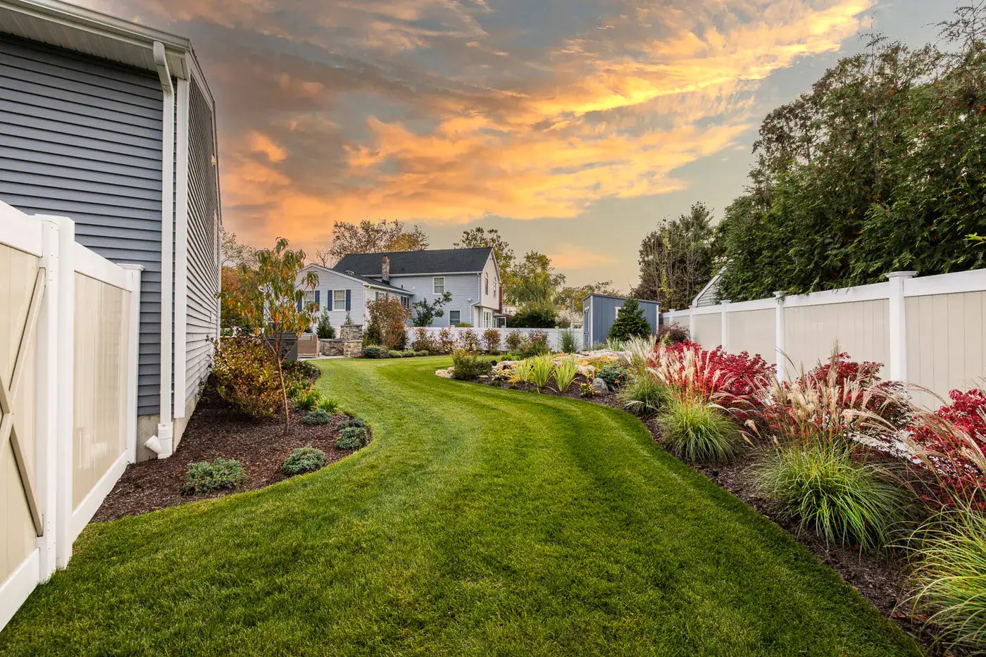 Modern backyard in New Jersey by CLC Landscape Design—paver patio with built‑in fire pit and cozy seating, overlooking a lush lawn edged with ornamental plantings under a vibrant sunset sky.