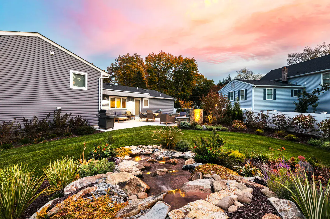 Contemporary backyard in New Jersey by CLC Landscape Design—paver patio patio with cozy seating under a soft sunset sky, lush lawn, and a flowing pondless stream with natural stone edging, creating a seamless fire‑and‑water focal point.