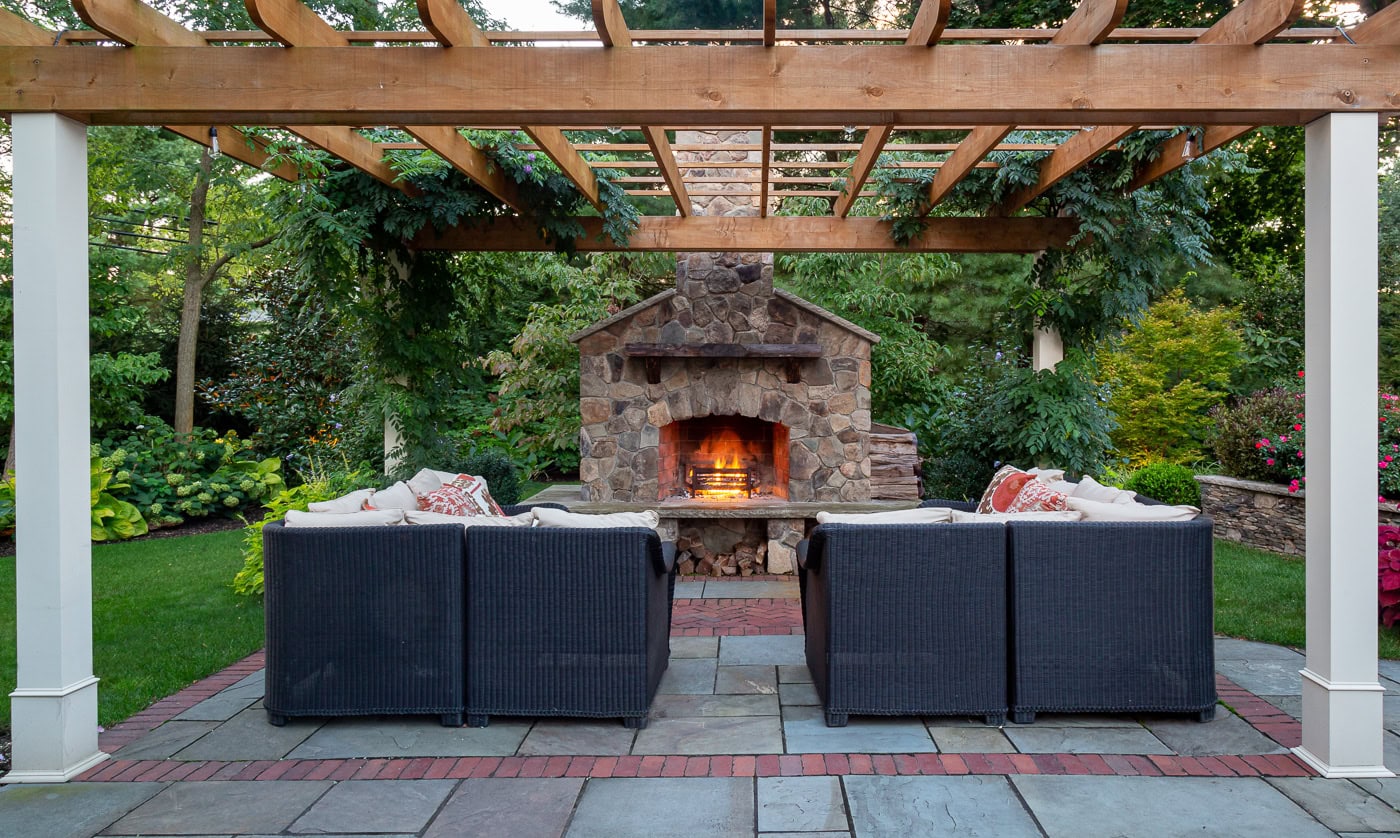 Stone outdoor fireplace surrounded by lush landscaping and colorful perennials in a Franklin Lakes backyard retreat designed by CLC Landscape Design.